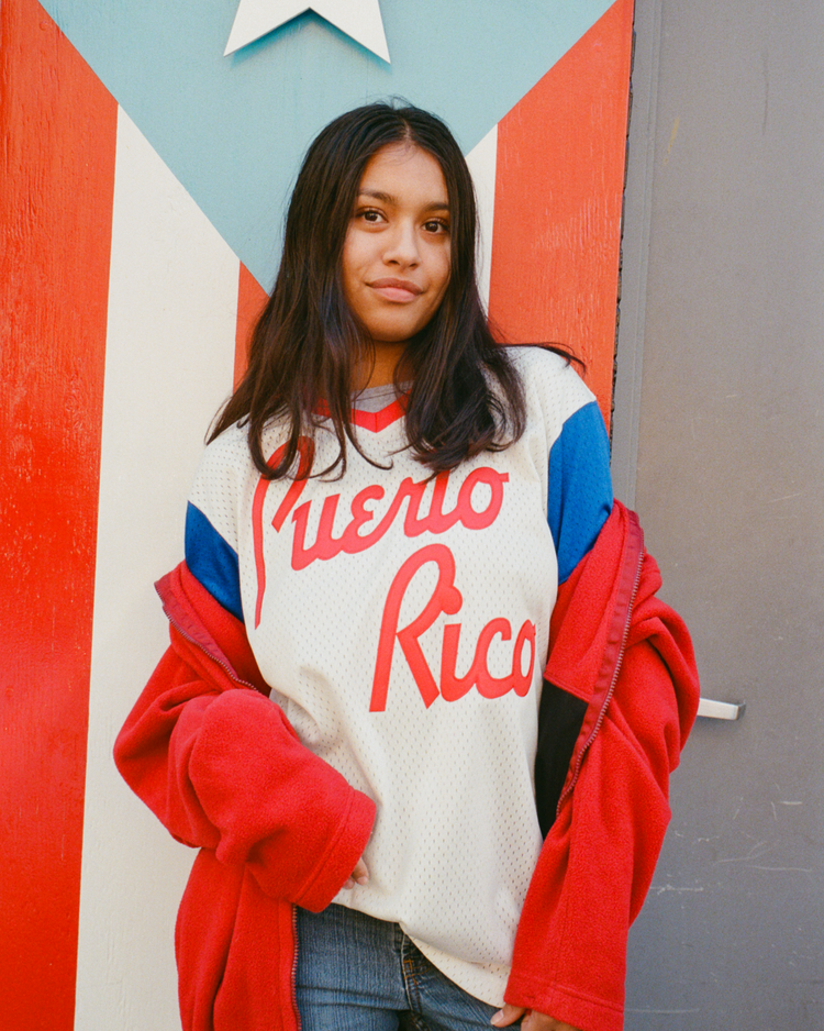 A woman with long dark hair wears an Ebbets Field Puerto Rico 1976 National Team Mesh V-Neck Jersey and a red jacket, standing before a wall painted like the Puerto Rican flag. She faces the camera with a slight smile.