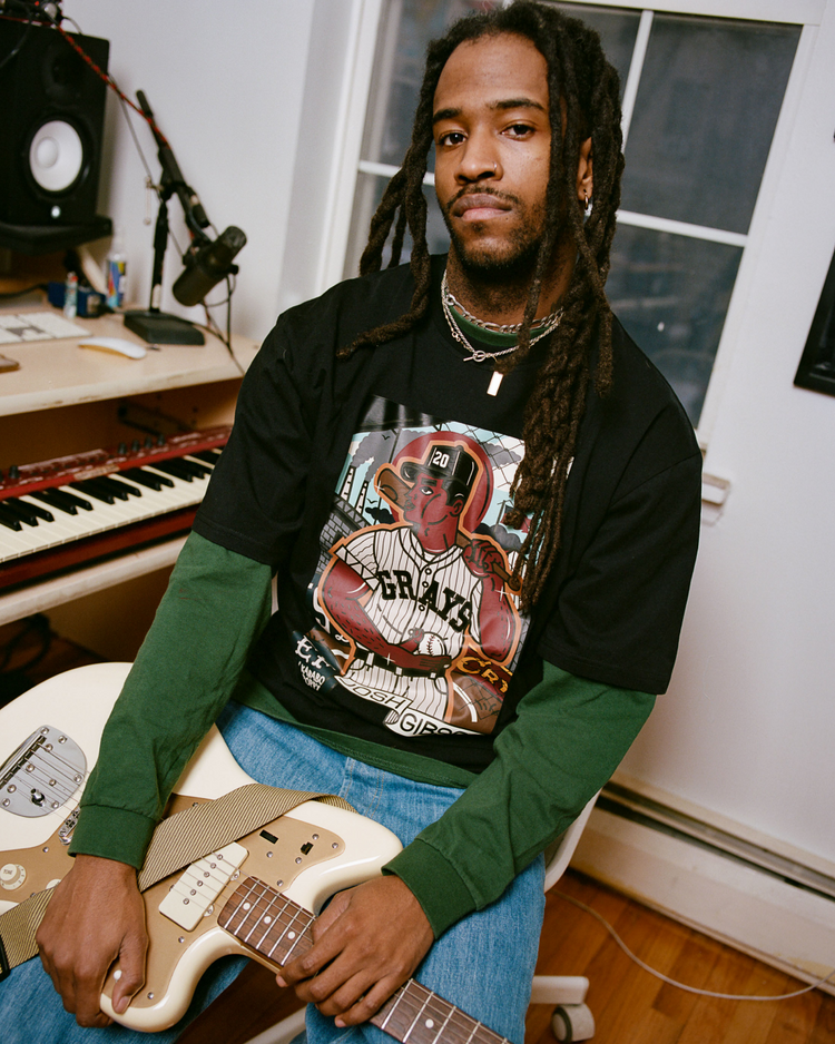 A man with long dreadlocks sits in a music studio holding a white electric guitar, wearing layered shirts and an Ebbets Field Karabo Poppy x Ebbets Field Josh Gibson Short Sleeve Graphic Tee; recording gear fills the background.