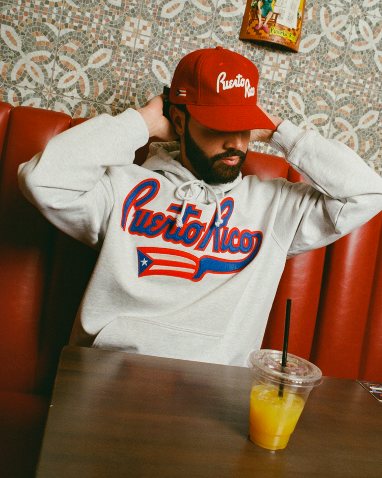 A man sits at a table with a drink, wearing an Ebbets Field Puerto Rico 1972 National Team Heavyweight Cotton Terry Pullover Hoodie.