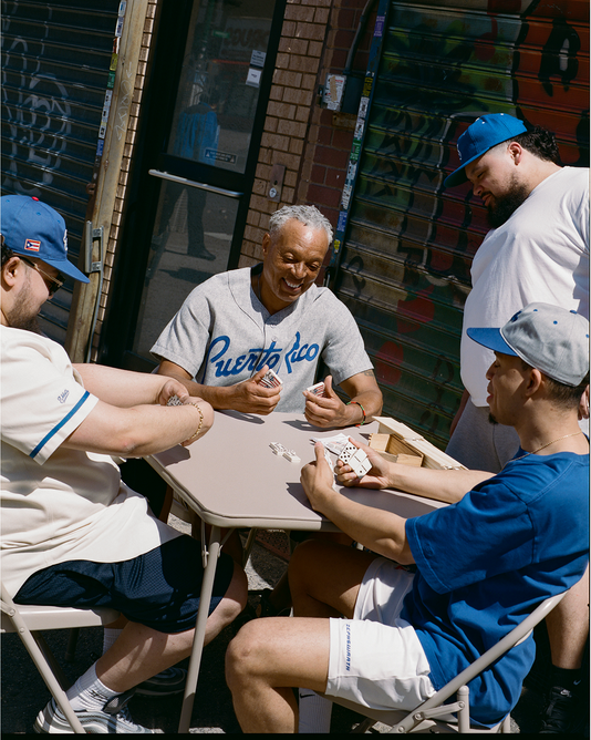 Four men sit around a small outdoor table playing cards, smiling and chatting. They wear casual clothes and caps. One man wears a Puerto Rico shirt. The setting is an urban street with a graffiti-covered metal door.