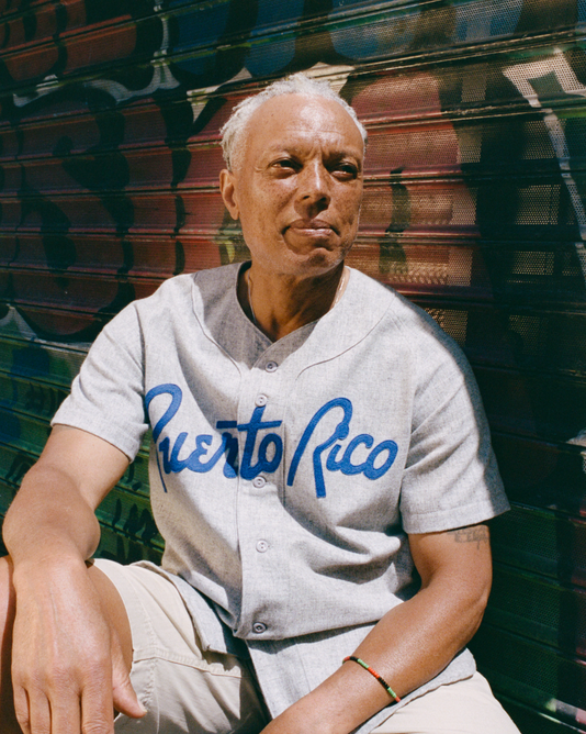 A person with short gray hair sits in front of a graffiti-covered metal gate, wearing a light gray Puerto Rico baseball jersey and light-colored shorts, looking off to the side in bright sunlight.