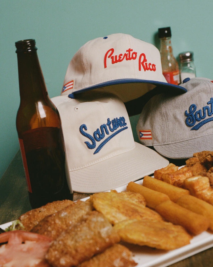Three baseball caps—including the Ebbets Field Santurce Cangrejeros 1939 Cotton Twill Ballcap—rest on a table with a beer bottle, hot sauce, and fried foods. Each cap features a small Puerto Rican flag and pops against the teal background.