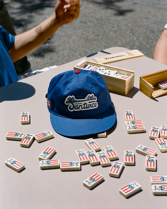 A blue Santurce baseball cap sits on a table scattered with dominoes featuring the Puerto Rican flag. An open wooden domino box and two people’s arms are visible in the background.