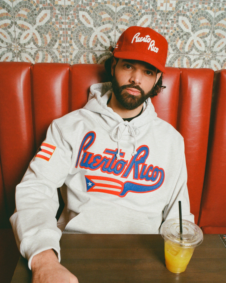 A bearded man in a gray hoodie and an Ebbets Field Puerto Rico 1958 National Team Wool Ballcap sits in a red booth with a yellow drink on the table, against a patterned tile wall.