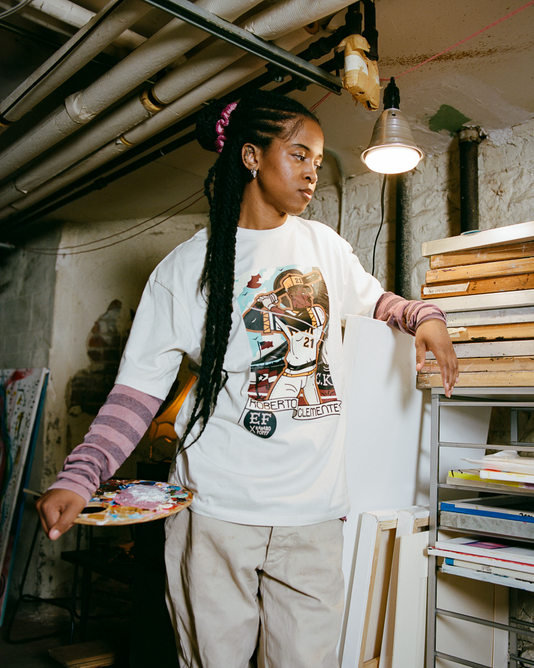 In an art studio, a person with long braids holds a paint palette and wears the Karabo Poppy x Ebbets Field Roberto Clemente Short Sleeve Graphic Tee by Ebbets Field, surrounded by supplies and canvases in celebration of Black History Month.
