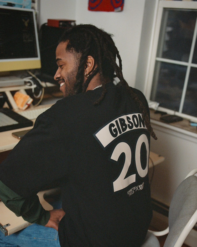A man with long dreadlocks smiles at his desk, wearing the Ebbets Field Karabo Poppy x Ebbets Field Josh Gibson Short Sleeve Graphic Tee, honoring Negro Leagues legend Josh Gibson amid computer equipment.
