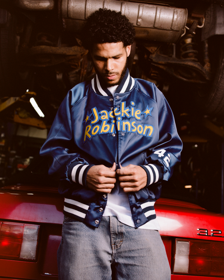 A man stands in a garage before a red car, buttoning his blue Ebbets Field Jackie Robinson Comic Book Hero Cotton Lined Satin Varsity Jacket and jeans. The car lift and undercarriage are visible overhead.
