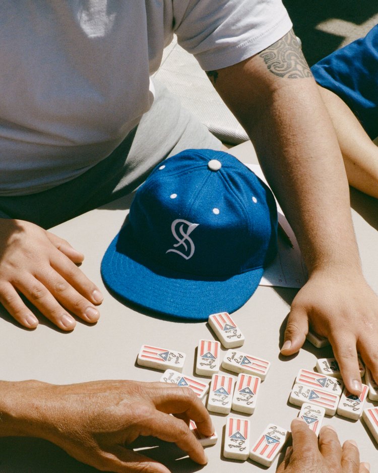 Two people play dominoes at a table, with an EFF Santurce Cangrejeros 1954 Authentic Wool Ballcap in blue resting nearby. One has a tattooed arm and both reach for American flag domino tiles.