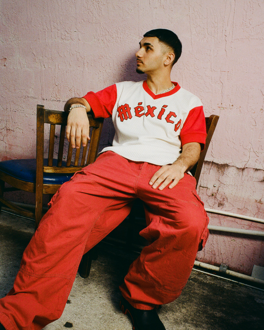 A young man wearing an Ebbets Field Diablos Rojos de Mexico V-Neck Mesh Jersey and baggy red pants sits on a wooden chair against a textured pink wall.