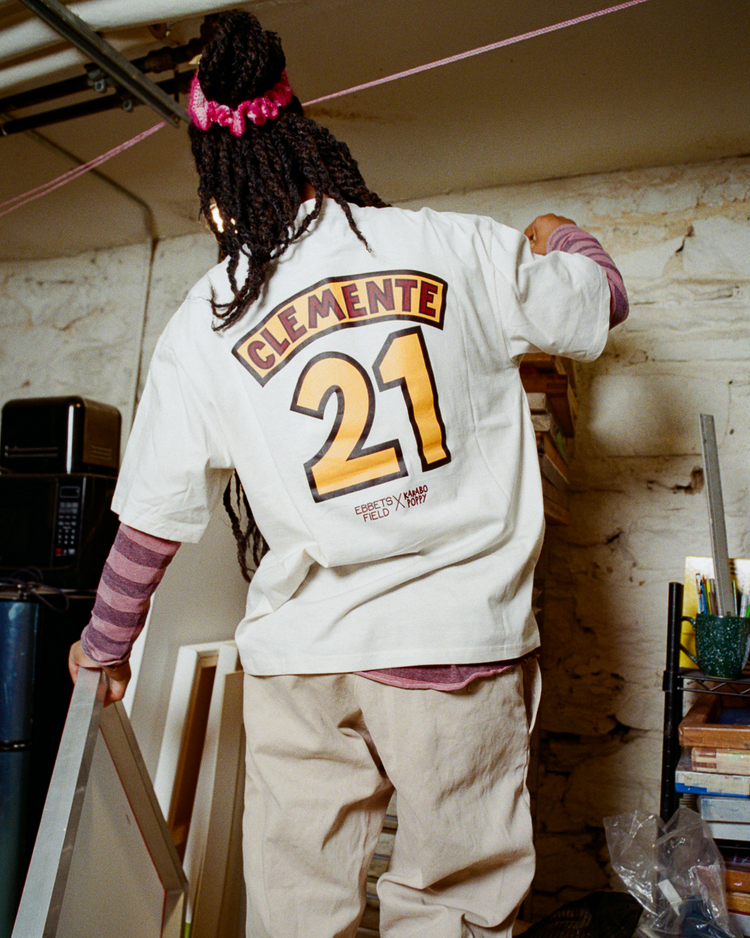 Wearing the Karabo Poppy x Ebbets Field Roberto Clemente Short Sleeve Graphic Tee by Ebbets Field, a person with long braids and a pink hairband stands with a picture frame in a room—an artistic, casual vibe ideal for Black History Month.