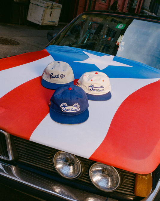 Three baseball caps reading Puerto Rico and Santurce rest on the hood of a car painted with the Puerto Rican flag, parked on a city street.