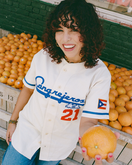 A smiling person with curly hair holds an orange toward the camera, standing in front of a fruit stand filled with oranges. They wear a white baseball jersey with Cangrejeros and a Puerto Rican flag patch.