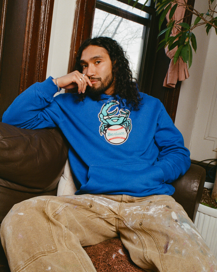 Wearing an Ebbets Field Santurce Cangrejeros Heavyweight Cotton Pullover Hoodie in bright blue, a person with long curly hair sits on a brown couch, paint-splattered pants on, with a plant and window in the background.