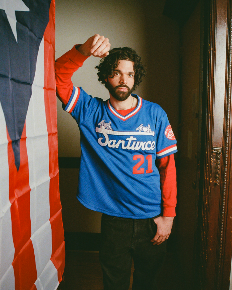 A man with curly hair and a beard wears an Ebbets Field Santurce Cangrejeros Mesh V-Neck Jersey, standing indoors by a large Puerto Rican flag, honoring the legacy of Puerto Rican baseball and Roberto Clemente.