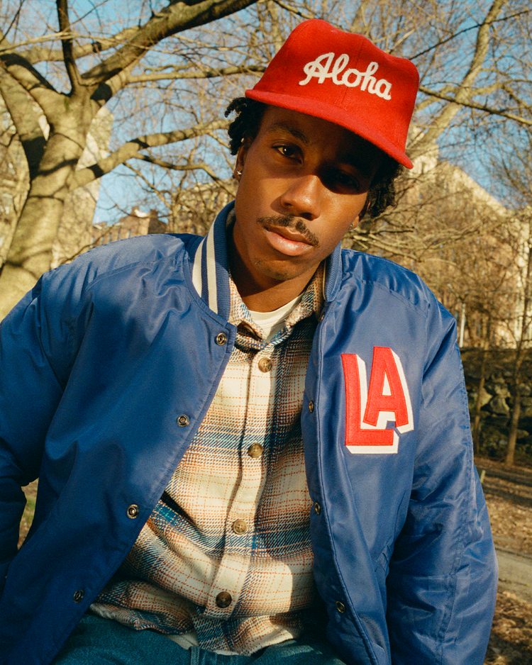 A person wearing a red Aloha cap, a plaid shirt, and an Ebbets Field Los Angeles Angels (PCL) Satin Varsity Jacket poses outdoors in a sunny park with leafless trees in the background.