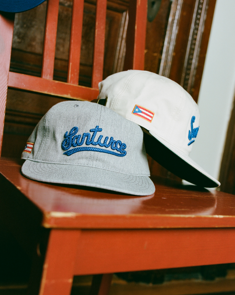Two Ebbets Field Santurce Cangrejeros 1939 Cotton Twill Ballcaps, in grey and white, sit on a red wooden chair, each embroidered with “Santurce” in a vintage style and finished with a small Puerto Rican flag patch.