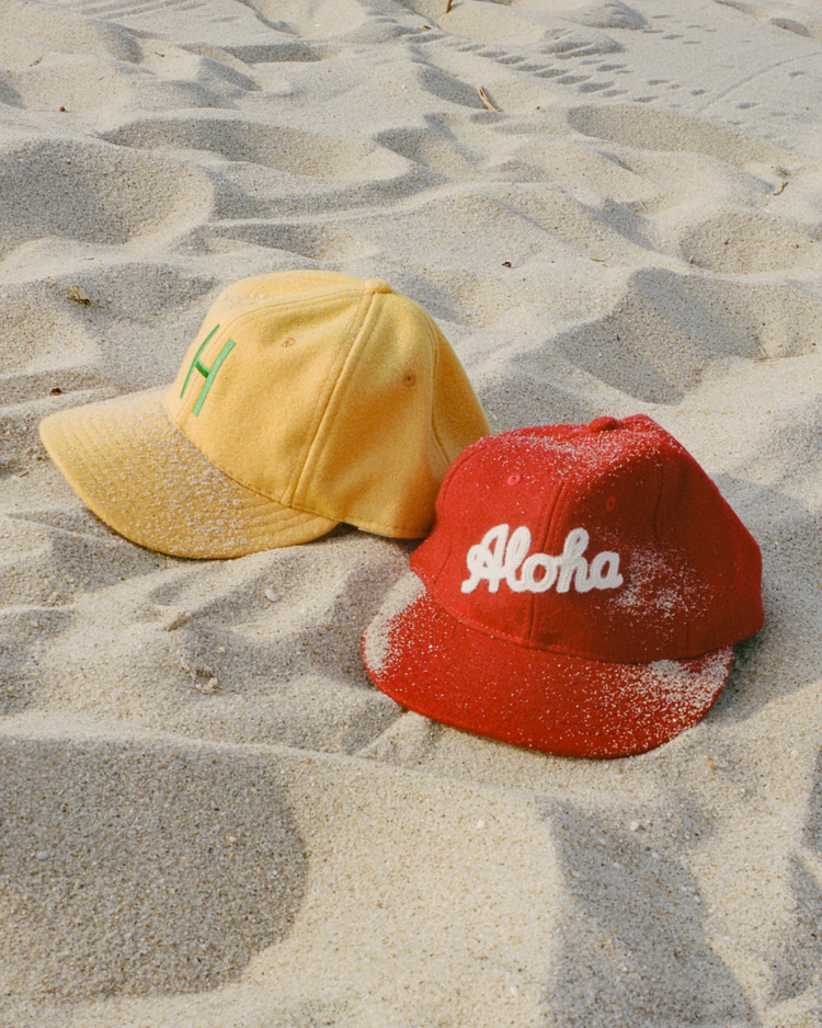 On the sandy beach, two EFF wool ballcaps rest—one yellow with a green H, and one red featuring the Aloha 100th Infantry 1942 logo—nods to Japanese-American history, both lightly sprinkled with sand.