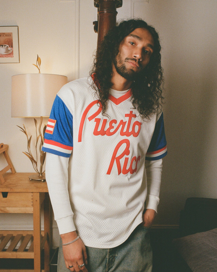 A bearded person with long curly hair stands indoors by a wooden side table and lamp, wearing the Ebbets Field Puerto Rico 1976 National Team Mesh V-Neck Jersey with blue and red accents.