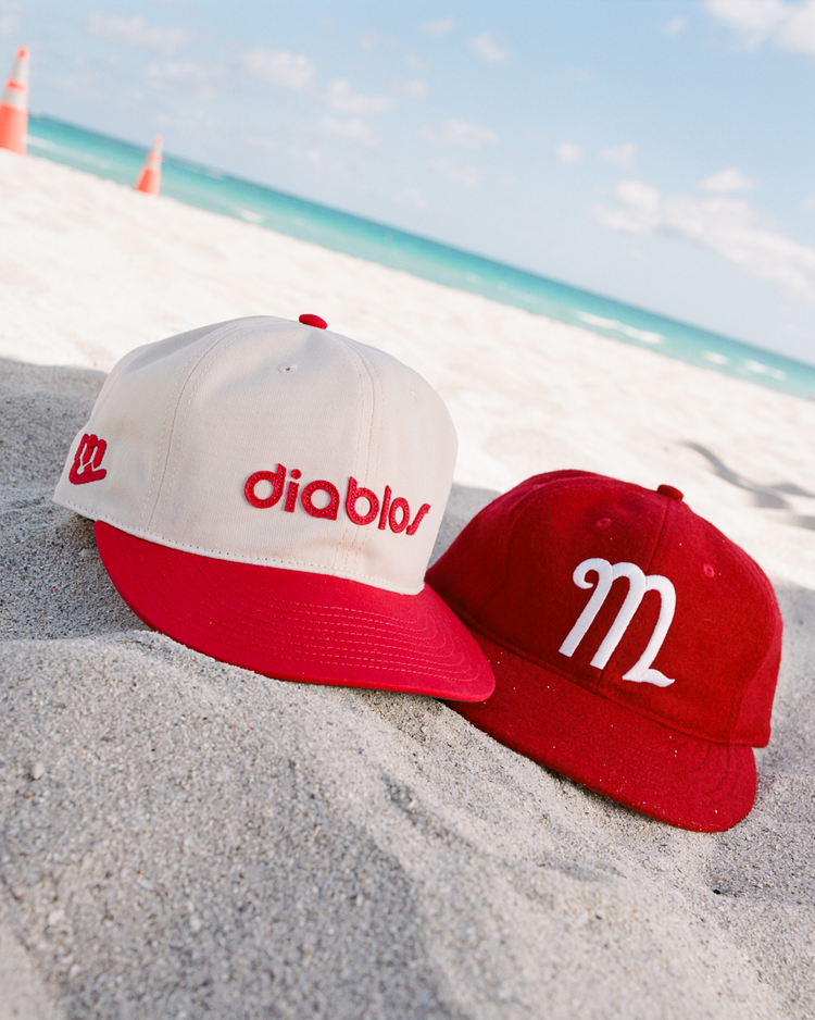 Two red-and-white Ebbets Field hats, including the Mexico City Diablos Rojos 1957 Authentic Wool Ballcap, sit on sandy beach under a blue sky. One cap reads “diablos,” the other has a stylized “M.” Orange cones are by the water.