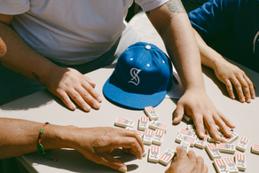 Four people sit around a table, reaching toward dominoes decorated with Puerto Rican flags and other symbols. A blue baseball cap is on the table, and one shirt reads Puerto.