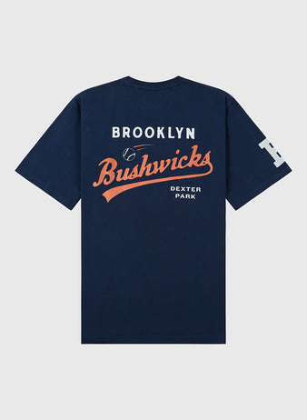 The Brooklyn Bushwicks Ebbets Tee by Ebbets Field is a navy blue T-shirt featuring white and orange Brooklyn Bushwicks and Dexter Park print on the back, a small baseball graphic, and a large white B on the left sleeve.