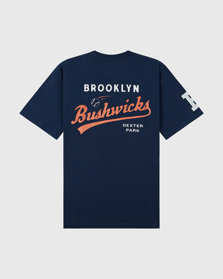 The Brooklyn Bushwicks Ebbets Tee by Ebbets Field is a navy blue T-shirt featuring white and orange Brooklyn Bushwicks and Dexter Park print on the back, a small baseball graphic, and a large white B on the left sleeve.