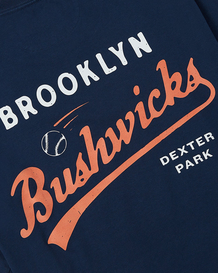 The Brooklyn Bushwicks Ebbets Tee by Ebbets Field features bold white and orange Brooklyn Bushwicks text, a flying baseball graphic, and Dexter Park printed in white on the lower right.