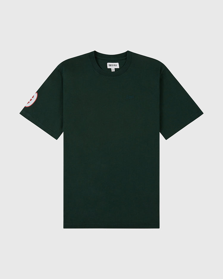 The Oakland Oaks Ebbets Tee with Back Print by Ebbets Field is a plain dark green short-sleeve t-shirt featuring a small round red and white patch on the left sleeve, shown against a light gray background.