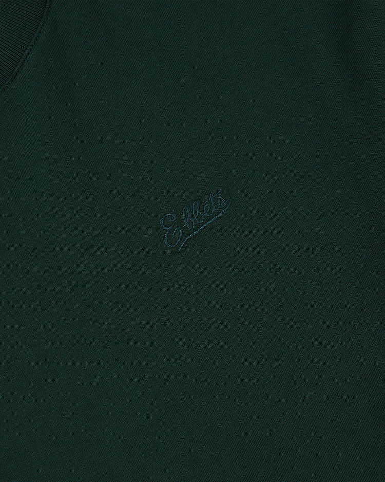 A close-up of dark green fabric from the Ebbets Field Oakland Oaks Ebbets Tee with Back Print, featuring Effets embroidered in matching thread in a small, cursive font at the center.