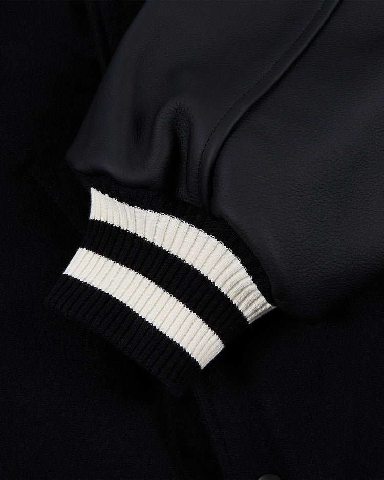 A close-up of the Harvard University Wool and Leather Varsity Jacket by Ebbets Field shows a black sleeve with a ribbed knit cuff featuring black and white stripes, crafted from leather or faux leather.