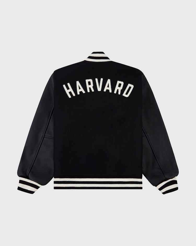 The Ebbets Field HARVARD VARSITY JACKET BLK features white striped cuffs, collar, and hem, with HARVARD in bold white letters on the back. Displayed on a plain light gray background.
