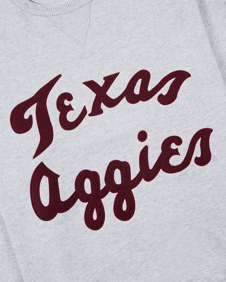 The Ebbets Field TEXAS A&M CREWNECK SS H-GRY is a gray sweatshirt featuring Texas Aggies in bold maroon cursive across the chest.