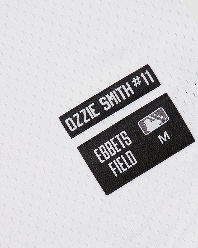 Close-up of a white Ozzie Smith Walla Walla Padres Mesh V-Neck Jersey with two black labels: one honoring the St. Louis Cardinals legend with his name and #11, the other showing Ebbets Field, MLB logo, and an M for medium size.