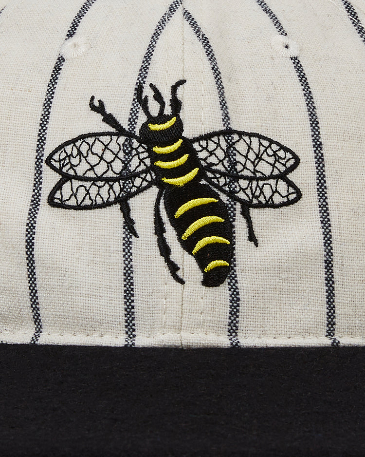 Close-up of the EFF Chicago Cats 1976 Authentic Wool Ballcap: beige fabric with vertical black pinstripes, a detailed yellow-and-black embroidered bee, and the edge of a black brim—echoing classic vintage soccer style.