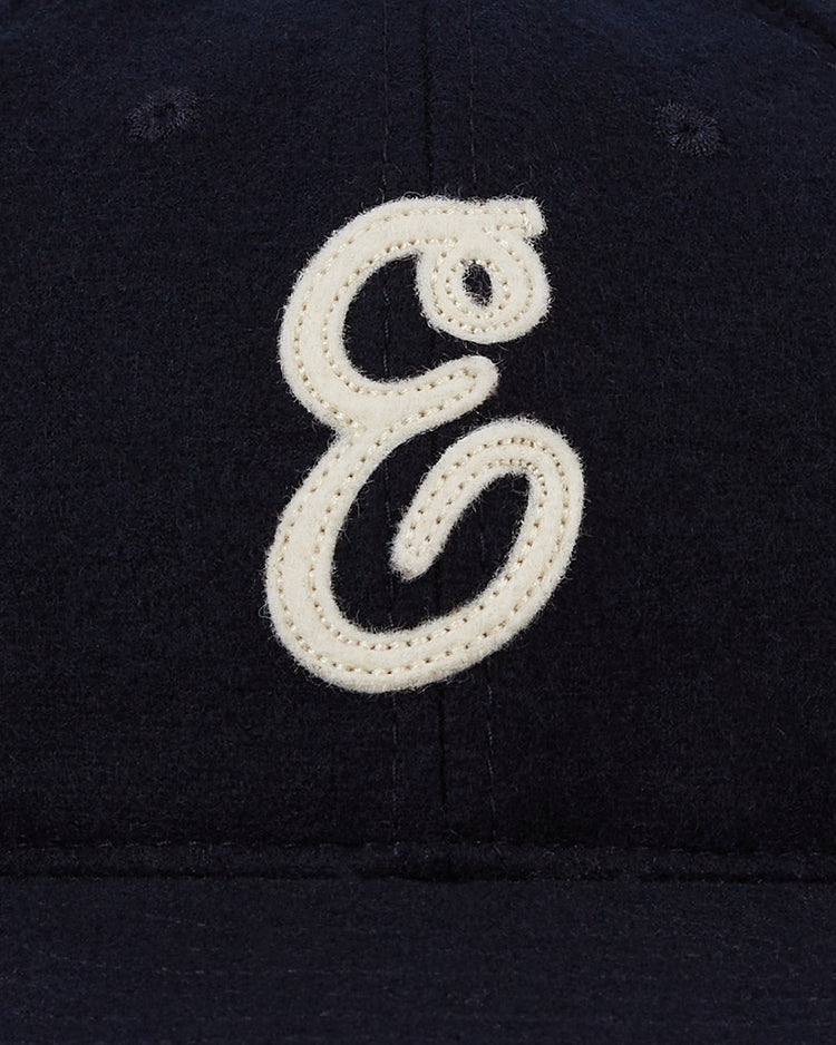 A close-up of the Ebbets Field Ebbets Felt ‘E’ Wool Ballcap, a dark wool vintage cap featuring a large white cursive embroidered “E” on the front.