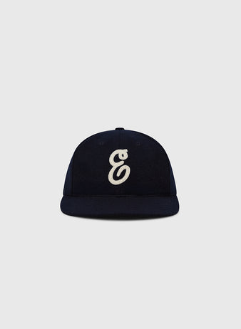 The Ebbets Felt 'E' Wool Ballcap by Ebbets Field is a navy wool broadcloth vintage cap with a white embroidered stylized 'E' on the front, shown against a light grey backdrop.