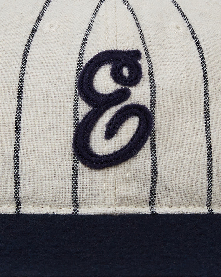 A close-up of the Ebbets Field Ebbets Felt 'E' Wool Pinstripe Ballcap, crafted from beige wool broadcloth with thin black pinstripes, a navy embroidered letter 'E' on the front, and a dark blue brim.