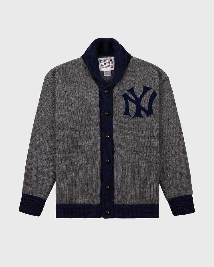 The Ebbets Field New York Yankees 1922 Wool Shawl Collar Sweater is a gray button-up with a dark blue collar, cuffs, hem, Yankees chest logo, and two front pockets, inspired by the team’s authentic 1922 sweater.