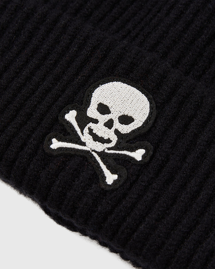 A black ribbed acrylic beanie by Ebbets Field, featuring an embroidered white skull and crossbones patch, inspired by the Sioux City Ghosts. Product: Sioux City Ghosts Embroidered Felt Ribbed Beanie.