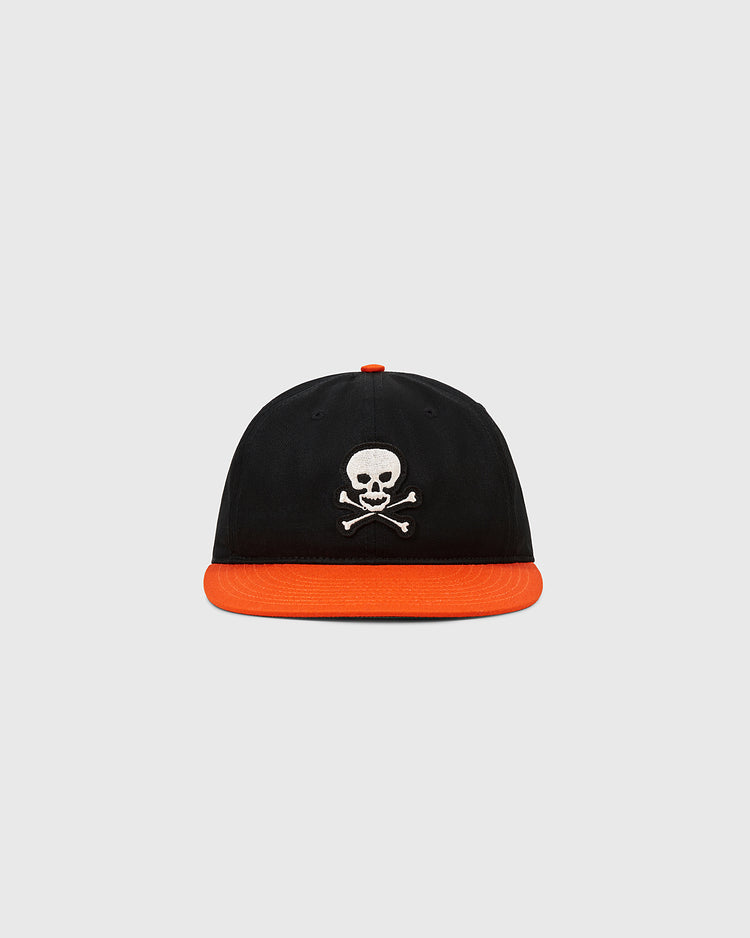 The Ebbets Field Sioux City Ghosts Cotton Twill Ballcap features a black crown, orange brim, and white skull and crossbones logo, inspired by the fast-pitch softball team. Displayed against a plain gray background.
