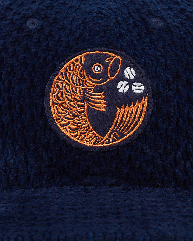 The Hiroshima Carp Japanese Indigo Corduroy Ballcap by Ebbets Field is a navy hat with textured fabric and an orange koi fish jumping over three coffee beans, embroidered in a circle.