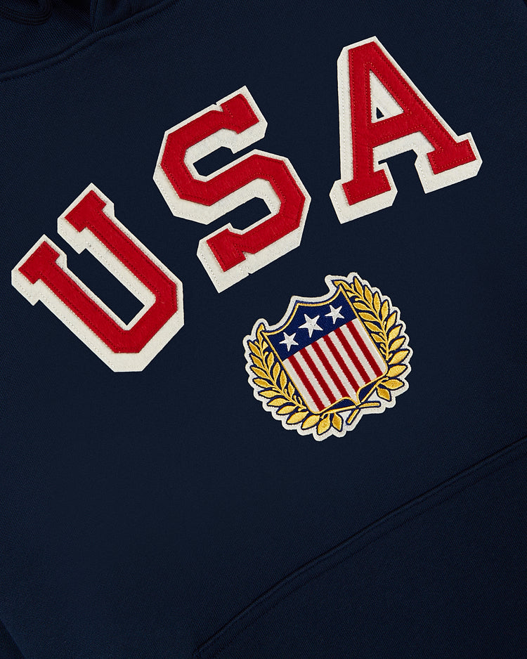Show your American pride in the USA 1960 Miracle On Ice Heavyweight Cotton Terry Pullover Hoodie by Ebbets Field, featuring navy blue fabric, bold red and white USA letters, and an embroidered shield with stars, stripes, and a gold laurel wreath.