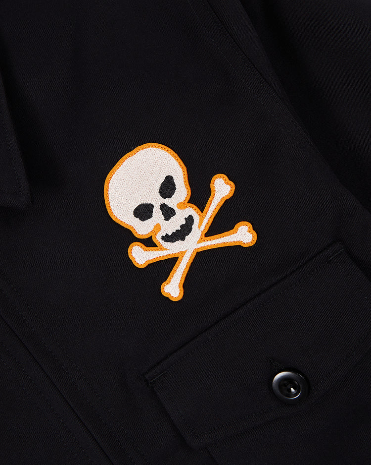 A close-up of the Ebbets Field Sioux City Ghosts Cotton Grounds Crew Jacket shows an embroidered skull and crossbones patch outlined in orange above a buttoned pocket, echoing vintage souvenir styles.