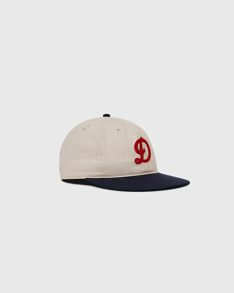 The Detroit Stars Cotton Twill Ballcap by Ebbets Field features a beige crown, navy blue brim, and a red embroidered “D” on the front, inspired by the 1920 Detroit Stars and shown against a plain light gray background.