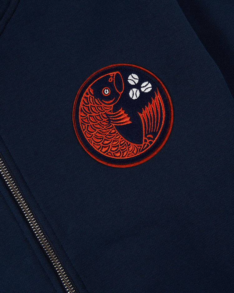 A close-up of the Ebbets Field Hiroshima Carp Heavyweight Cotton Terry Full-Zip Hoodie in navy, featuring a red embroidered fish patch and three white shapes beside a silver zipper.