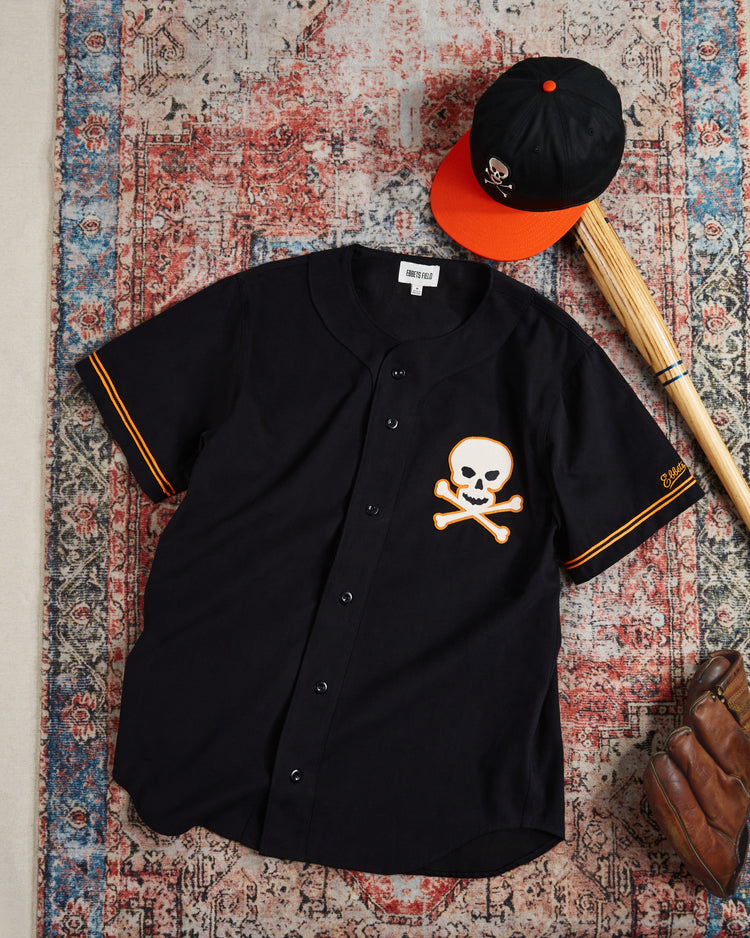 The Ebbets Field Sioux City Ghosts Cotton Twill Baseball Jersey, featuring a vintage skull and crossbones logo, pairs with a matching black cap—an ideal set for fast-pitch softball fans. Displayed on a patterned rug with a wooden bat.