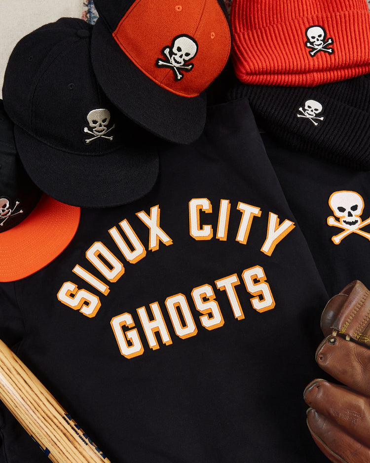 The Ebbets Field Sioux City Ghosts Cotton Grounds Crew Jacket—classic black with orange lettering—is displayed among skull-logo caps, beanies, a glove, and a bat: a vintage must-have for fast-pitch softball collectors.