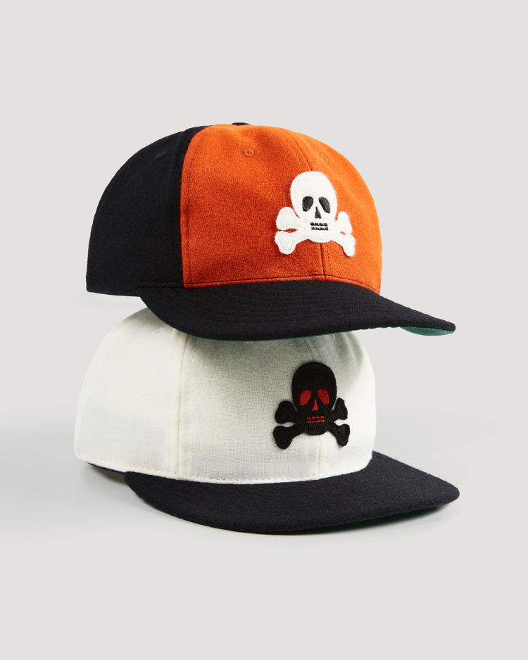 Two baseball caps: on top, an orange and black Sioux City Ghosts 1935 Authentic Wool Softbrim Ballcap from EFF with a white skull and crossbones; below, a vintage-style white and black ballcap with a black skull featuring red eyes and mouth.