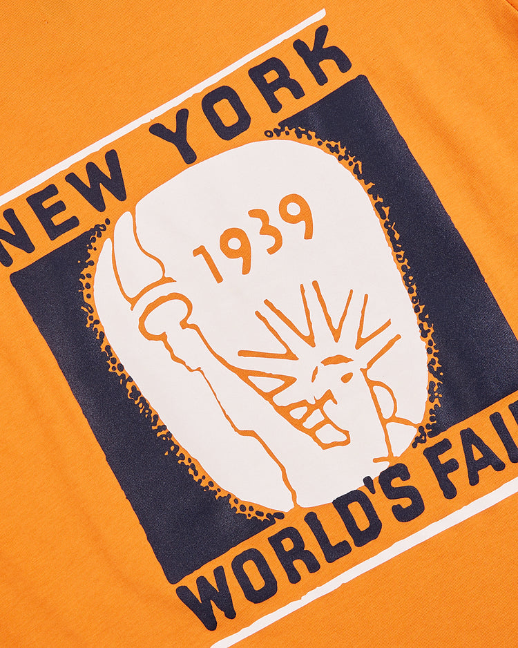 The Ebbets Field New York 1939 World’s Fair Short Sleeve Graphic Tee features bold graphics of the Statue of Liberty and event text on orange, ideal for Yankees apparel fans or anyone wanting a classic fair look.