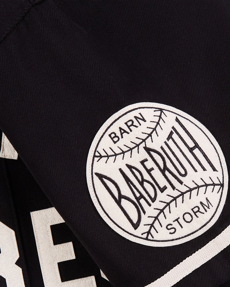 Close-up of a black fabric patch on the Ebbets Field Babe Ruth Bustin' Babes Cotton Twill Baseball Jersey, featuring a white baseball graphic and bold BARN, BABE RUTH, and STORM lettering inspired by 1927 barnstorming tours.